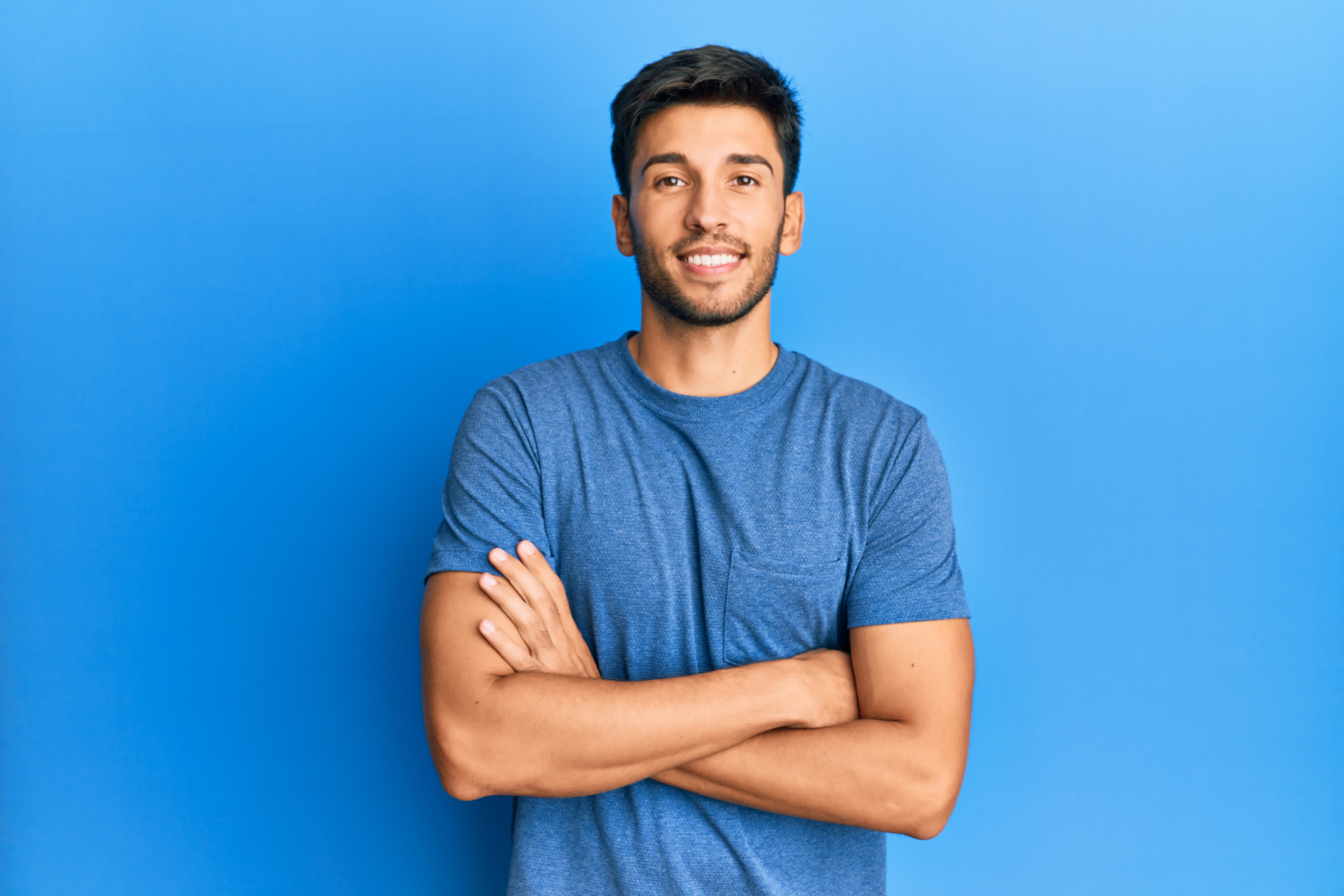 joven hombre guapo con camiseta informal sobre fondo azul cara feliz sonriendo con los brazos cruzados mirando la camara persona positiva